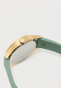 Analogové hodinky se zlatým pouzdrem, zeleným silikonovým páskem a lesklým povrchem. Mají kulatý ciferník a korunku pro nastavení.