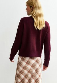 Maglione a maglia bordeaux con vestibilità morbida e polsini a costine, abbinato a una gonna a quadri beige con tonalità marroni e ruggine.