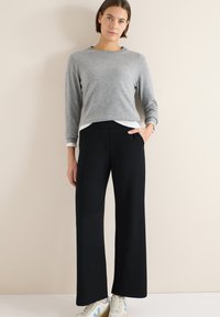 Femme portant un pull gris à manches longues, un pantalon noir à jambes larges et des baskets blanches, debout contre un fond beige avec une main dans la poche.
