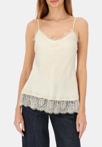 Jasnokremowa koszulka na ramiączkach z koronkową lamówką u dołu i przy dekolcie. Regulowane ramiączka, miękki materiał oraz luźny fason z fakturowanymi koronkowymi akcentami.