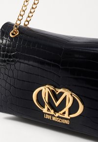 Borsa a tracolla in pelle verniciata nera con motivo coccodrillo, tracolla in catena dorata e logo dorato in evidenza con le lettere "M" intrecciate e "LOVE MOSCHINO."