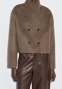 Brown wool double-breasted jacket with a high collar, featuring two black buttons. Paired with brown leather pants, displaying a textured finish.