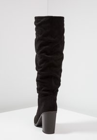 Bota de ante negro sobre la rodilla con un diseño arrugado, que cuenta con un tacón grueso y un tejido ligeramente texturizado. Se muestra la vista trasera.