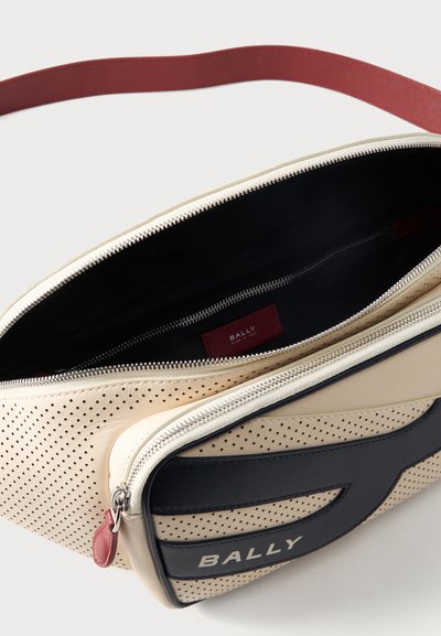 Τσαντάκι χιαστί από κρεμ διάτρητο δέρμα με μαύρη μπροστινή τσέπη, ασημένια φερμουάρ, κόκκινη ταινία τραβήγματος και κόκκινο ρυθμιζόμενο λουράκι, ανοιχτό για να δείχνει το εσωτερικό.