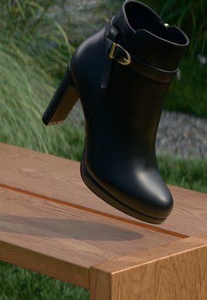 Black leather ankle boot with a rounded toe and gold hardware accents, featuring a strap around the ankle. Set against a wooden surface.