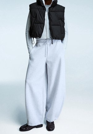 Personne portant un pantalon de jogging large gris clair, un haut à fermeture éclair gris clair, une doudoune courte noire et des chaussures marron foncé.