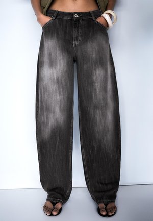 Osoba nosiaca voľné, vyblednuté čierne rifle s rukami v vreckách a čierne sandále s otvorenou špičkou na svetlom jednofarebnom pozadí.