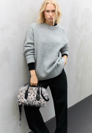 Blonde woman wearing a gray sweater and black pants, holding a small white and black spotted faux fur handbag, standing against a light gray wall.
