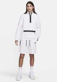 Balts vilnas džemperis ar ceturtdaļas rāvējslēdzēju priekšā, melns rāvējslēdzējs un kabatu akcenti, pārī ar baltām šortiem un Nike zīmolu uz kājas.