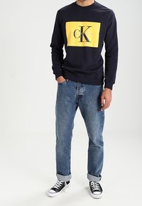 Sudadera azul marina con gráfico de caja amarilla, que presenta "Calvin Klein Jeans" y un gran logo "CK", combinada con jeans de lavado claro arremangados y zapatillas negras.