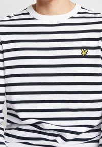 Maglione a righe con maniche lunghe in bianco e blu scuro, caratterizzato da collo e polsini a coste, con un piccolo logo giallo ricamato sul petto.