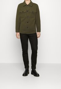 Olivengrøn uldblanding jakke med krave, to brystlommer og sorte knapper. Sammen med tætsiddende sorte bukser og sorte ankelstøvler.