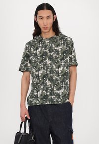 Tričko s krátkym rukávom s motívom zelených paliem na krémovom pozadí. Model drží čiernu tašku; tmavé denimové nohavice dopĺňajú outfit.