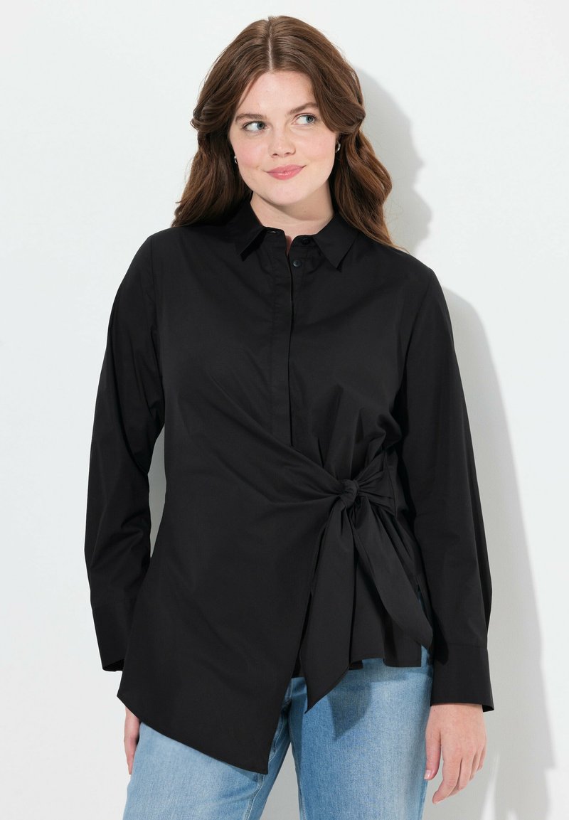 Blusa negra de manga larga, con cuello de botones y un detalle anudado en un lado. Hecha de tela suave, combinada con jeans azules.