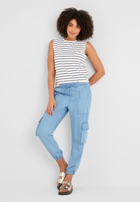 Woman wearing sleeveless white and black striped top, light blue cargo jogger pants, and metallic buckle sandals, standing against white wall.