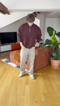 Felpa bordeaux con zip, jeans baggy grigi chiari e sneakers argentate. La stanza presenta un pavimento in legno, una pianta in vaso e mobili moderni.