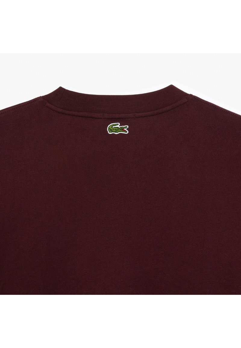 Chemise à manches longues bordeaux avec une texture côtelée et un col rond. Elle comporte un logo crocodile brodé vert à l'arrière.