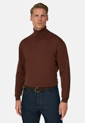Mand iført en tætsiddende brun rullekravebluse med lange ærmer og mørkeblå jeans med et flettet brunt bælte.