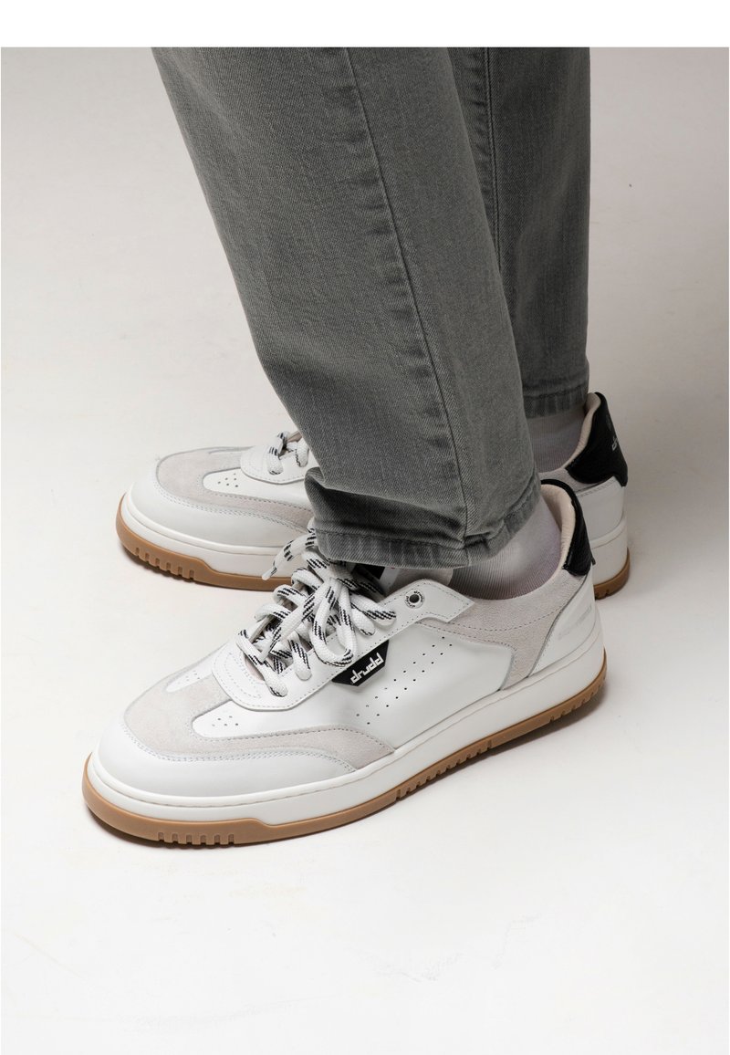 White and gray sneaker with suede and leather panels, black accents, lace-up closure, and a textured gum rubber sole. Designed for casual wear.