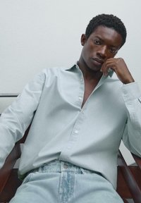 Un giovane con i capelli ricci e corti è seduto su una sedia indossando una camicia azzurro chiaro con bottoni e jeans azzurro chiaro, poggiando la testa sulla mano.