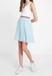 Lichtblauwe geplooide rok met een aansluitende tailleband met rode en marineblauwe strepen, gecombineerd met een witte geribde tanktop en witte sneakers.