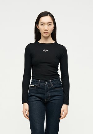 Γυναίκα με εφαρμοστό μαύρο πουλόβερ μακρυμάνικο με το λογότυπο "Guess Jeans" και σκούρο ψηλόμεσο τζιν, στέκεται μπροστά σε απλό ανοιχτόχρωμο φόντο.