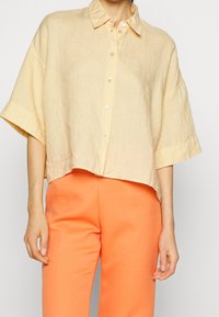 Kvinde iført en løs, kortærmet beige skjorte med knapper, parret med højtaljede, lyse orange bukser mod en ensfarvet baggrund.