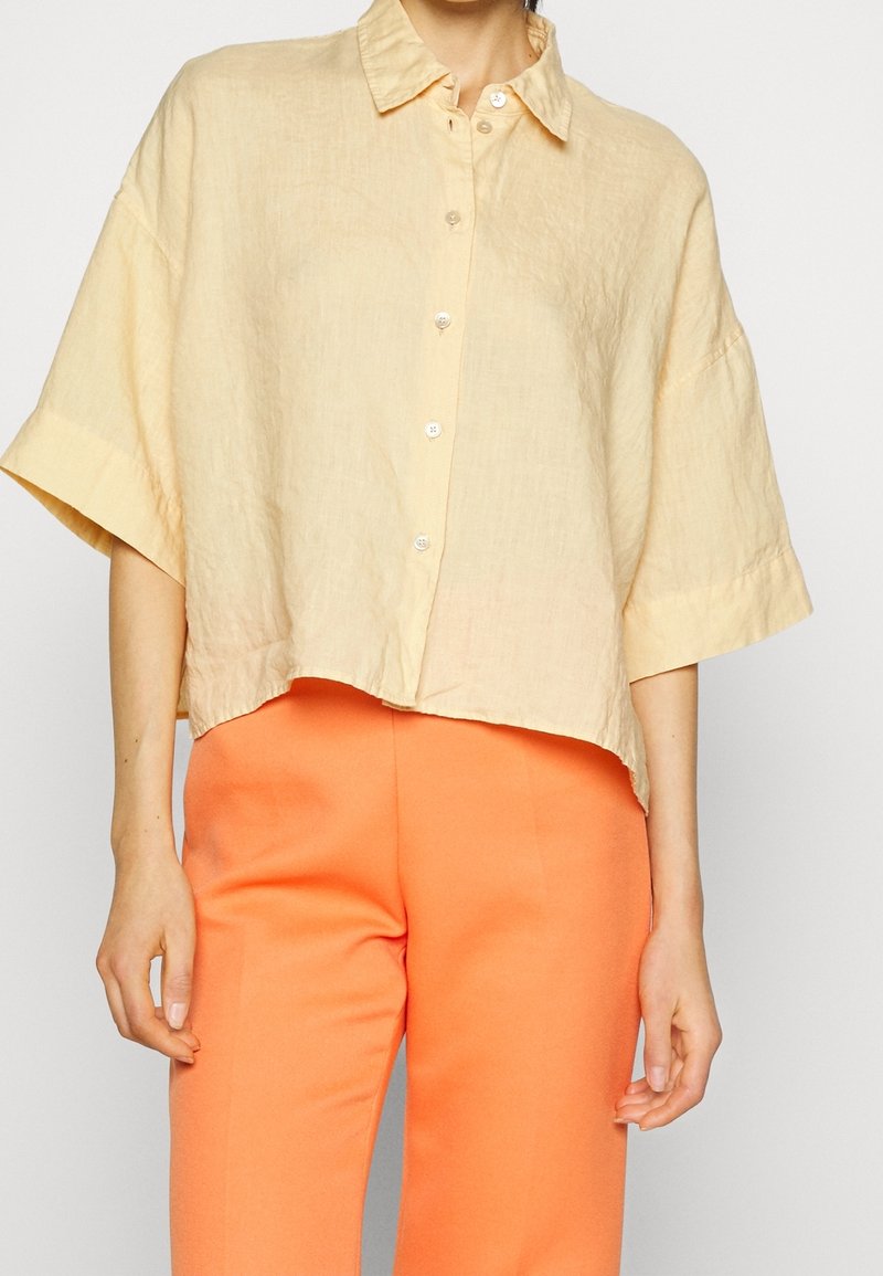 Kvinde iført en løs, kortærmet beige skjorte med knapper, parret med højtaljede, lyse orange bukser mod en ensfarvet baggrund.