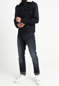 Granatowa kurtka z kołnierzem z owczej wełny, zapinana na guziki, z dwoma kieszeniami na piersiach, zestawiona z ciemnymi dżinsami i białymi trampkami.