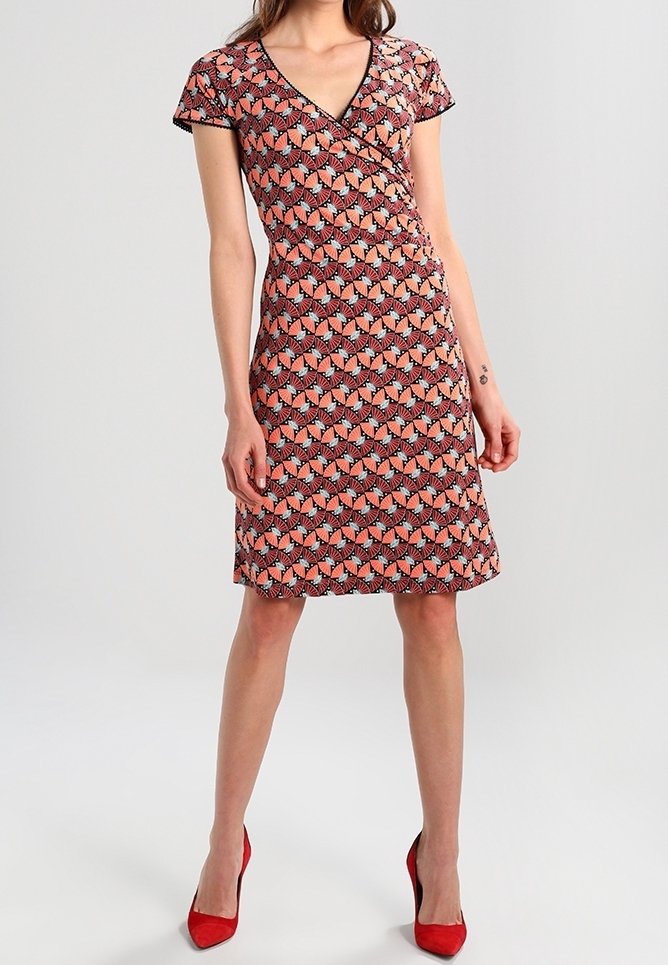 Červené a černé vzorované zavinovací šaty s krátkými rukávy a výstřihem do V. Tkanina je hladká. Kombinováno s červenými lodičkami na vysokém podpatku.