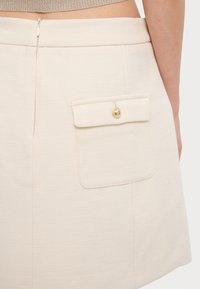 Jupe mini beige en tissu texturé, avec fermeture éclair au dos et poche à rabat ornée d'un bouton doré.
