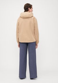 Beige kapucňová bunda s elastickými manžetami a voľným strihom, spojená so širokými nohavicami zloženými z texturovanej tmavomodrej látky.