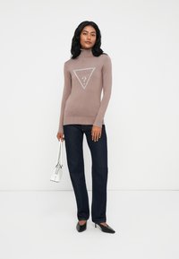 Donna che indossa un maglione a collo alto color malva con il logo "GUESS", jeans scuri, tacchi neri e tiene in mano una piccola borsa bianca, in piedi contro uno sfondo bianco.