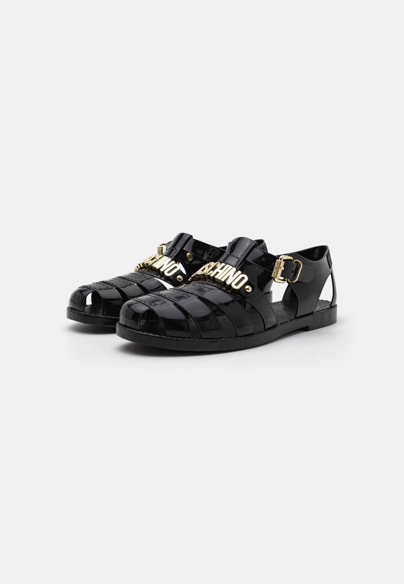 Sandales en caoutchouc noir avec un design entrecroisé, bout ouvert, quincaillerie dorée avec le lettrage "MOSCHINO" et un accent de boucle sur le côté.