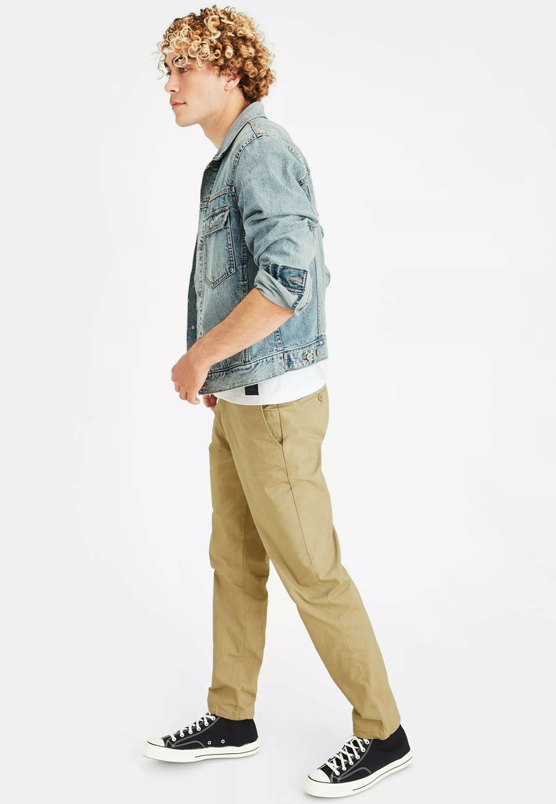 Jeansjasje met een lichte blauwe wassing, versleten patches op de mouwen, een wit T-shirt eronder, gecombineerd met tan kleurige broek en zwarte sneakers.
