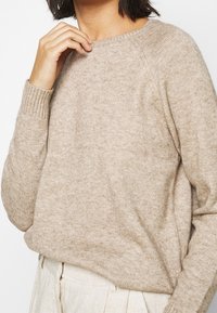 Pull en maille beige avec un col rond, des poignets et un ourlet côtelés. Texture douce avec une coupe décontractée, présenté avec un bas de ton neutre.