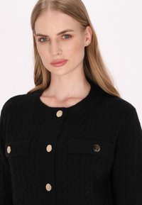 Mustat kabeliknit-neulecardigan kultasävyisillä napilla, pyöreä pääntie ja kaksi etutaskua. Pehmeä tekstuuri ja tiukkakulkuinen malli.