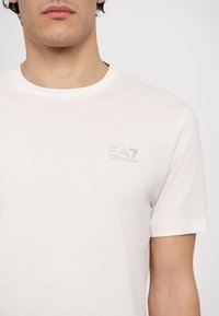 Biela bavlnená tričko s crew neckom s malým vyšitým logom EA7 v svetle šedej farbe, krátke rukávy a mäkká, textúrovaná tkanina.