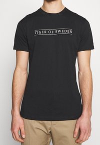 Muž v černém tričku s krátkým rukávem s nápisem "Tiger of Sweden" na hrudi a béžových kalhotách, zobrazený od krku po boky.