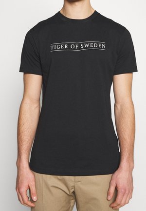 Hombre con una camiseta negra de manga corta que lleva el texto "Tiger of Sweden" en el pecho y pantalones beige, mostrado de cuello a caderas.
