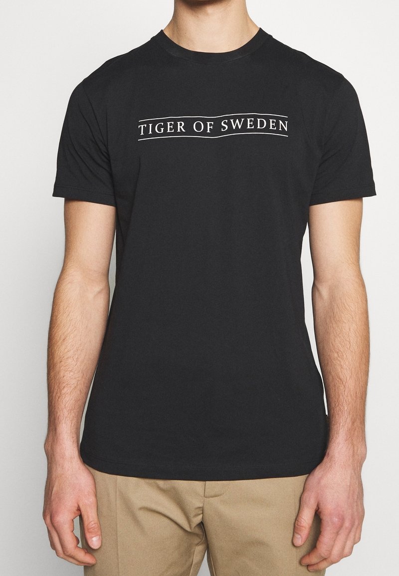 Muž v černém tričku s krátkým rukávem s nápisem "Tiger of Sweden" na hrudi a béžových kalhotách, zobrazený od krku po boky.