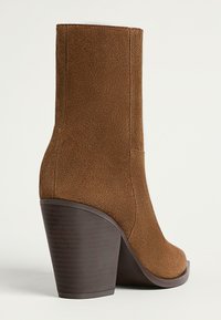 Stivaletto in suede marrone con tacco alto e sagomato, una finitura liscia e una silhouette affusolata. La suola è leggermente testurizzata.