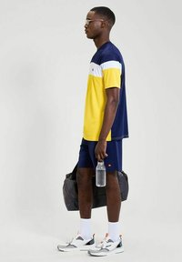 Camiseta deportiva de manga corta en amarillo, marino y blanco; combinada con pantalones cortos marinos. Los accesorios incluyen una bolsa gris y una botella de agua transparente. Calcetines blancos y zapatillas.