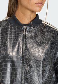 Crna bomber jakna s teksturom krokodila, zatvaranjem na rajfešlus, rebrastim ovratnikom i bijelim prugastim akcentima na rukavima. Suptilni detalj logotipa na prsima.