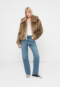 Giacca in pelliccia sintetica marrone con collo ampio, abbinata a un maglione leggero e jeans blu a gamba dritta. Sneakers nere completano l'outfit.