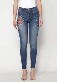 Smal passform mörkblå jeans med blommönster broderat på vänster lår och lätt slitage, bärs med klara remmar på klackarna.