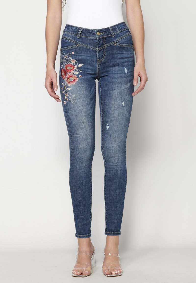 Smal passform mörkblå jeans med blommönster broderat på vänster lår och lätt slitage, bärs med klara remmar på klackarna.