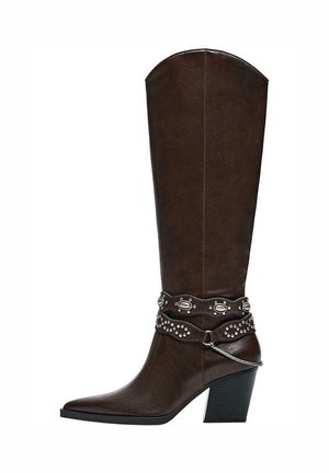 Tamno smeđa kožna čizma do koljena sa šiljastim vrhom, srednje visokom blok petom i ukrasnim remenjem sa zakovicama te srebrnim lancem oko gležnja.