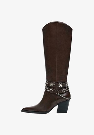 Tamno smeđa kožna čizma do koljena sa šiljastim vrhom, srednje visokom blok petom i ukrasnim remenjem sa zakovicama te srebrnim lancem oko gležnja.