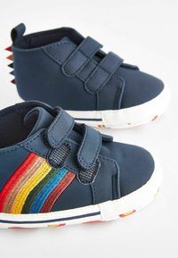 Chaussons bleu marine avec deux bandes Velcro. Présentent un design multicolore à rayures arc-en-ciel. Semelle en caoutchouc blanc avec des accents colorés sur le bord.